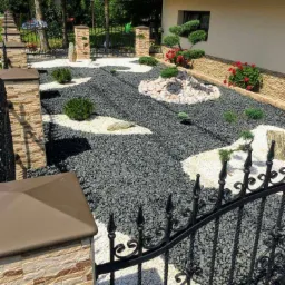 Ogród przydomowy z czarnym żwirem, białymi plamami z kamyków, ozdobnymi krzewami i elementami skalnymi, widziany zza ozdobnego, kutego ogrodzenia.