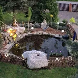 Oczko wodne z oświetleniem punktowym i kamienną dekoracją, otoczone zielenią i niskim płotkiem z wikliny, widok z góry, wieczorne oświetlenie.