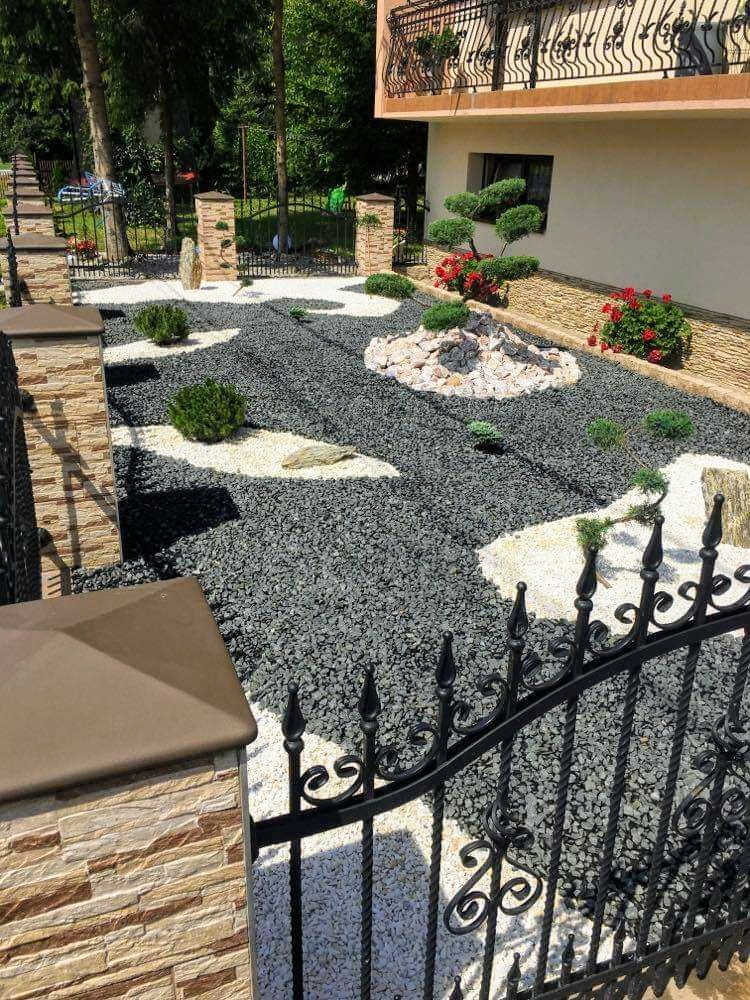 Ogród przydomowy z czarnym żwirem, białymi plamami z kamyków, ozdobnymi krzewami i elementami skalnymi, widziany zza ozdobnego, kutego ogrodzenia.