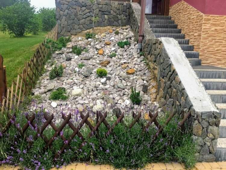 Skarpa z kamieni i roślinności zabezpieczona drewnianym płotkiem, prowadząca do kamiennych schodów wejściowych do domu.