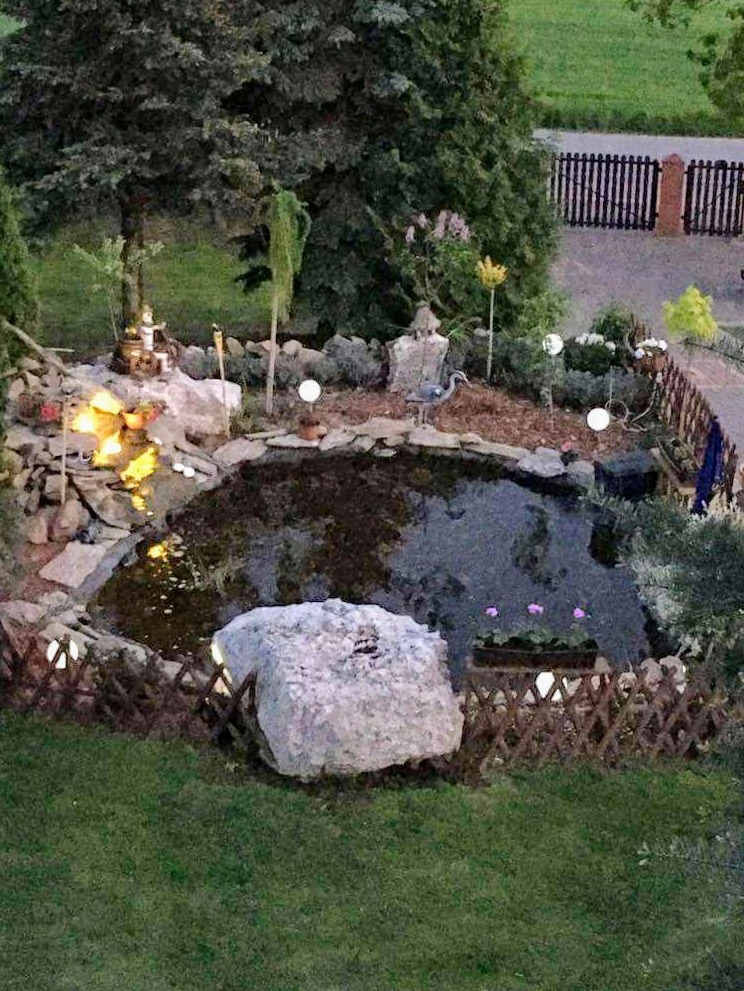 Oczko wodne z oświetleniem punktowym i kamienną dekoracją, otoczone zielenią i niskim płotkiem z wikliny, widok z góry, wieczorne oświetlenie.