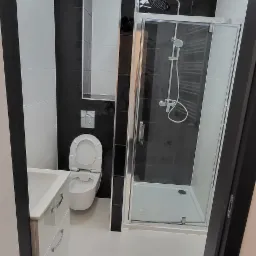 Nowoczesna łazienka z kabiną prysznicową ze szklanymi drzwiami, toaletą podwieszaną i umywalką z szafką, utrzymana w biało-czarnej kolorystyce z akcentami czarnych płytek na ścianach i podłodze.