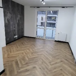 Pokój z nową podłogą z paneli ułożonych w jodełkę, szara ściana akcentująca, czarne listwy przypodłogowe, oświetlenie szynowe na suficie, para butów i ubrania leżące na podłodze.