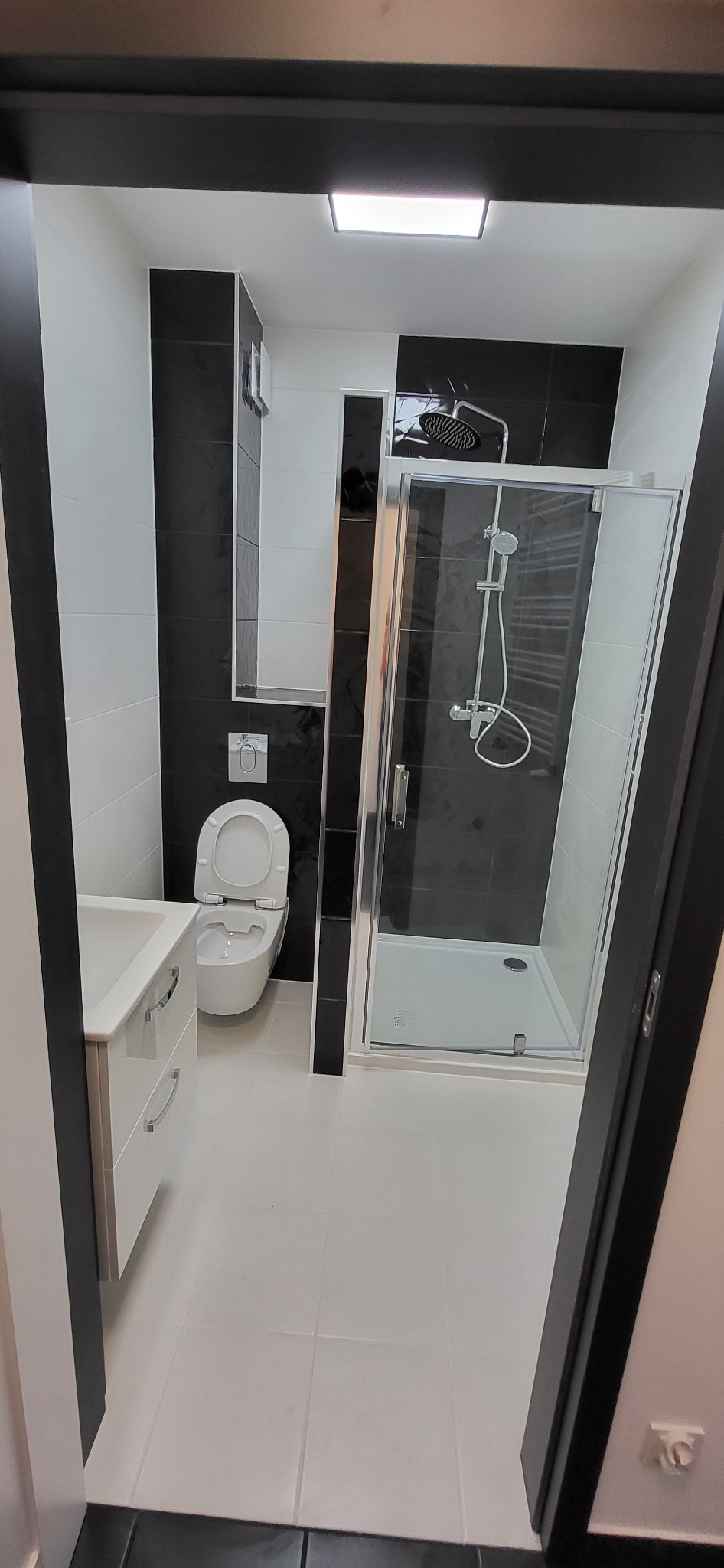 Nowoczesna łazienka z kabiną prysznicową ze szklanymi drzwiami, toaletą podwieszaną i umywalką z szafką, utrzymana w biało-czarnej kolorystyce z akcentami czarnych płytek na ścianach i podłodze.