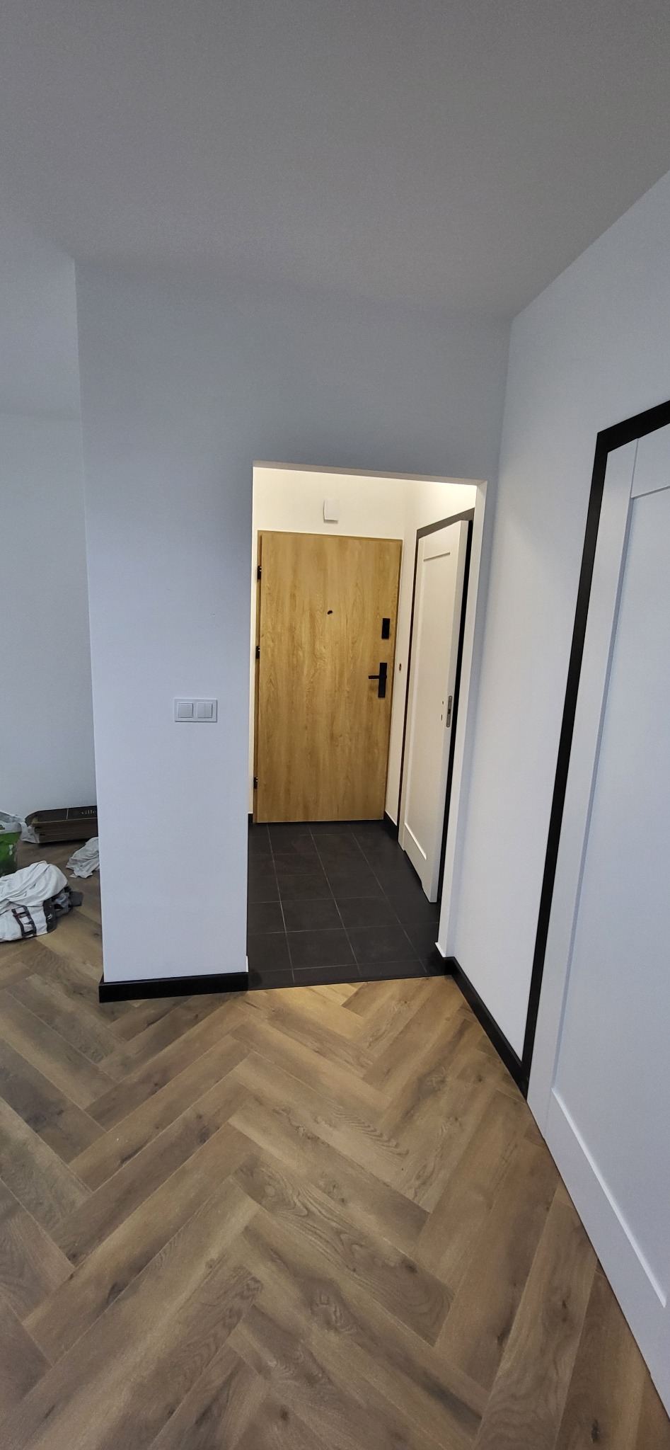 Ułożona podłoga z paneli w jodełkę, widok na przedpokój z czarną podłogą z płytek i drzwi wejściowe.