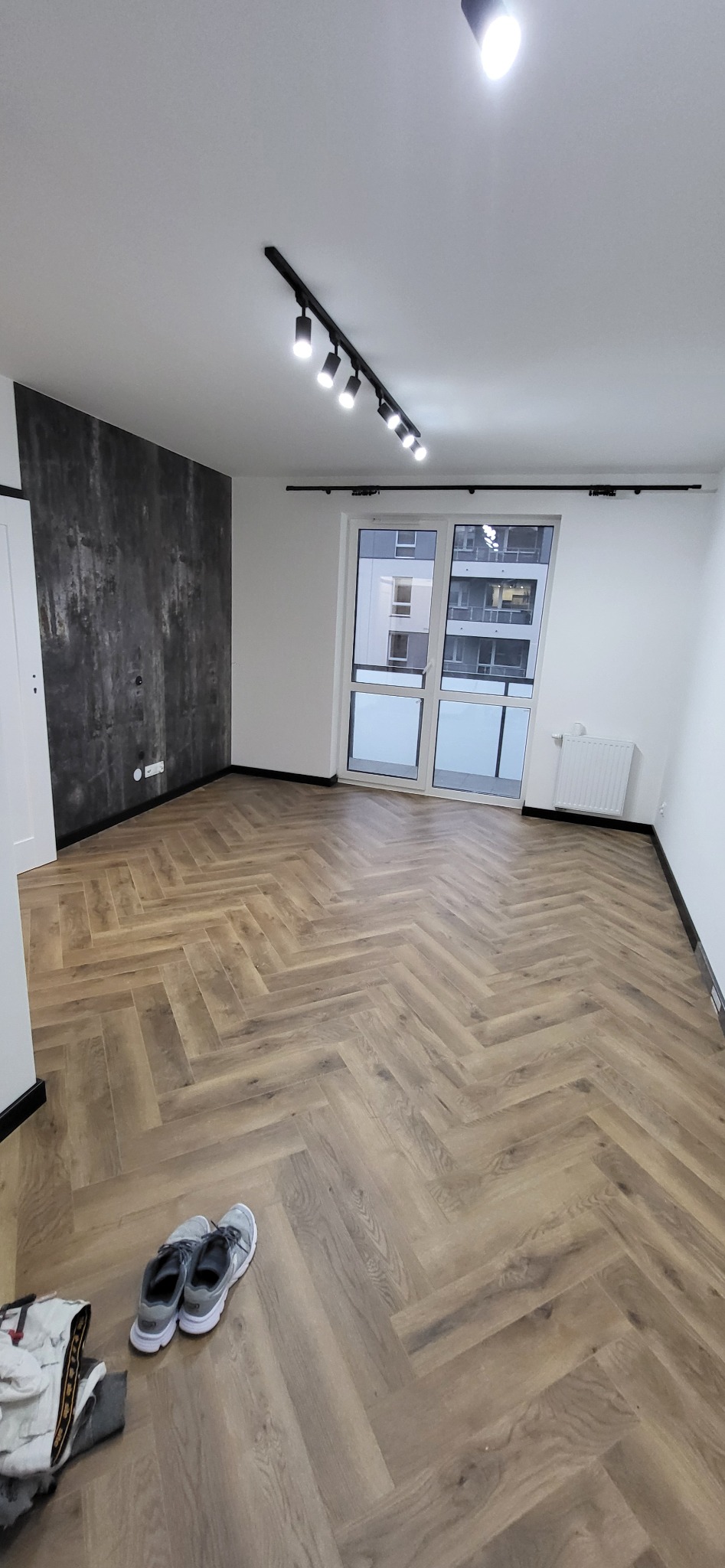 Pokój z nową podłogą z paneli ułożonych w jodełkę, szara ściana akcentująca, czarne listwy przypodłogowe, oświetlenie szynowe na suficie, para butów i ubrania leżące na podłodze.