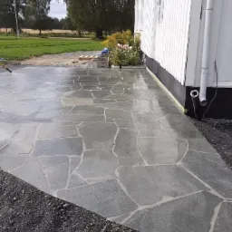 Świeżo ułożona, nieregularna nawierzchnia z kamienia naturalnego, spryskiwana wodą, przylegająca do białej ściany budynku z widocznymi rynnami i rabatą kwiatową w tle.