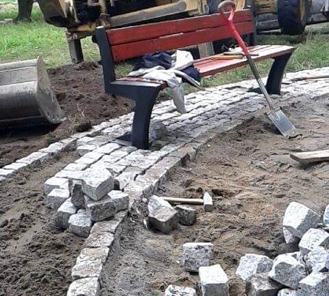 Fragment ścieżki z kostki granitowej w trakcie budowy, z ławką parkową w tle, widoczna łopata i młotek brukarski.