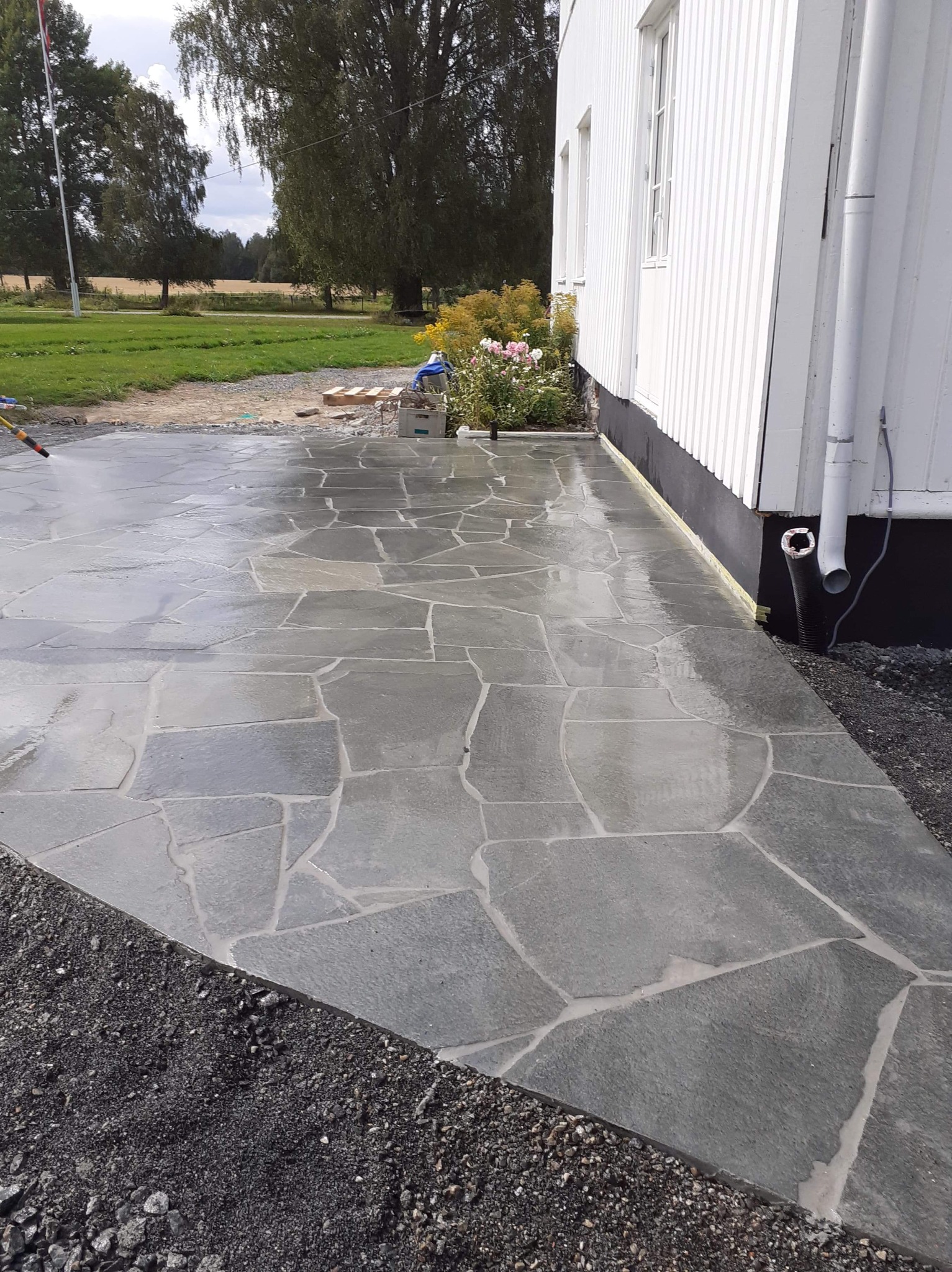 Świeżo ułożona, nieregularna nawierzchnia z kamienia naturalnego, spryskiwana wodą, przylegająca do białej ściany budynku z widocznymi rynnami i rabatą kwiatową w tle.