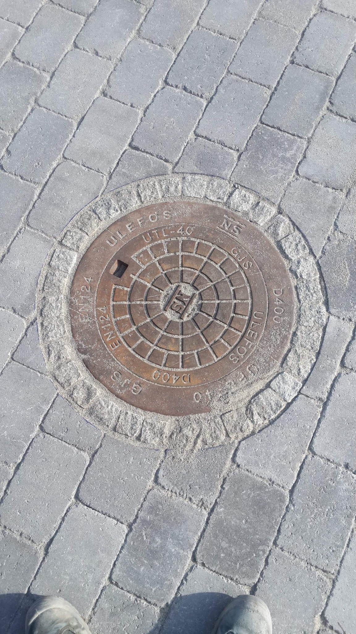 Szare prostokątne kostki brukowe z okrągłą metalową studzienką kanalizacyjną ULEFOS UTL-40 GJS D400 EN124 z widocznym fragmentem buta w dolnej części kadru.