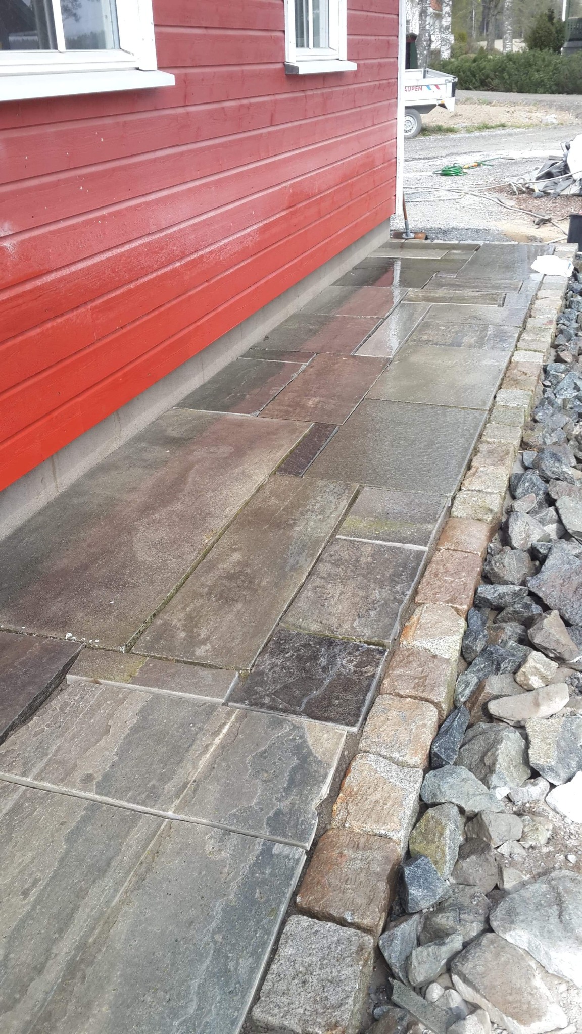 Ułożona ścieżka z kamiennych płyt obok czerwonej ściany budynku, z obrzeżem z kostki granitowej i podsypką z kamieni.