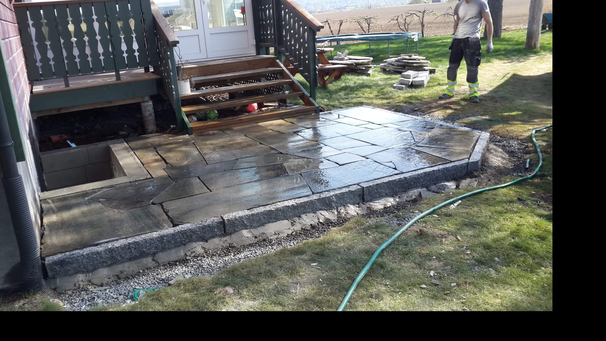 Nieregularne, mokre płyty kamienne tworzą patio przy schodach domu, obrzeża z szarego granitu, widoczny wąż ogrodowy i fragment trawnika.