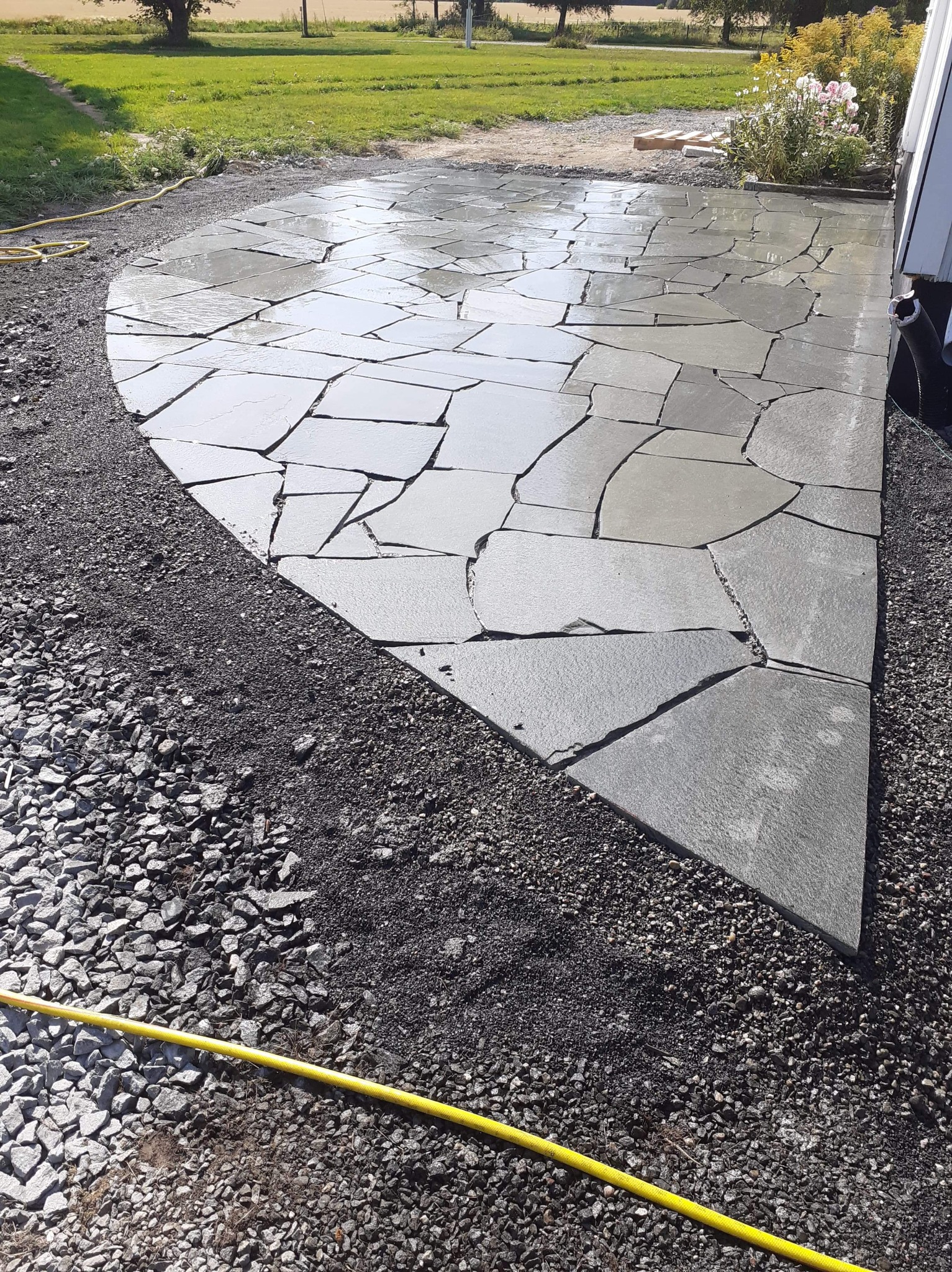 Nieregularny kształt podłoża wyłożony szarą płytą kamienną, otoczony ciemnym żwirem, z widocznym fragmentem żółtego węża ogrodowego i zielonym trawnikiem w tle.