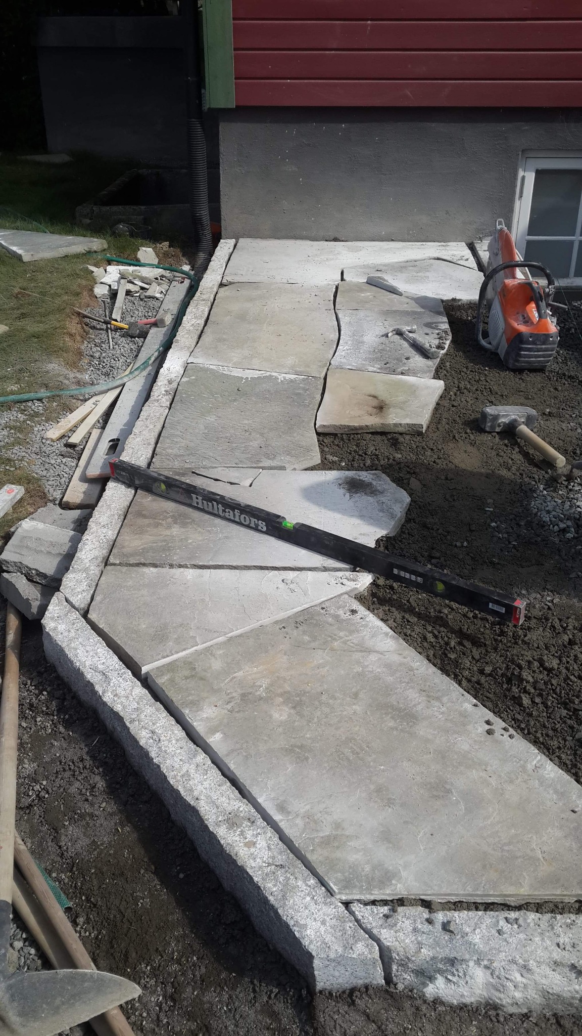 Fragment układanej ścieżki z nieregularnych płyt kamiennych, obramowanej szarą kostką granitową, z poziomicą Hultafors, piłą do kamienia i młotkiem na ziemi, na tle ściany budynku z oknem w piwnicy.