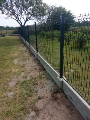 Długie ujęcie zainstalowanego czarnego ogrodzenia z siatki panelowej z betonową podmurówką w ogrodzie, widoczne słupki i drzewa w tle.