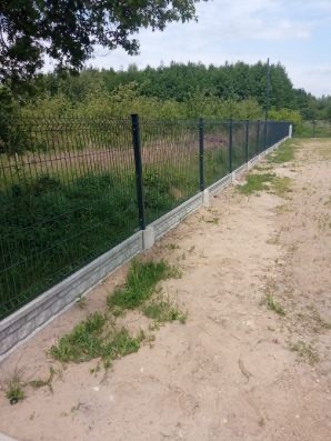 Długie, proste ogrodzenie z paneli z podmurówką betonową na piaszczystym gruncie, z widokiem na zieleń w tle.