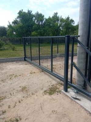 Przesuwna brama z paneli z siatki, szara, widoczna na tle zieleni, zamontowana przy betonowym słupie.