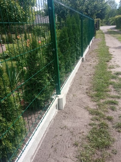 Zielone ogrodzenie panelowe na betonowej podmurówce, widoczne słupki i siatka, w tle drzewa i ścieżka.