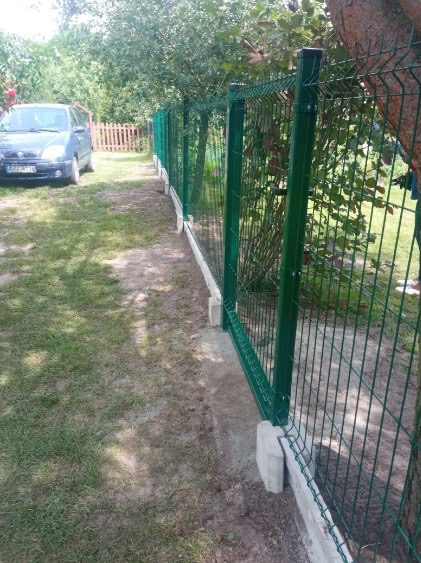 Zielone panele ogrodzeniowe z siatki z betonową podmurówką, widok z boku na tle trawnika i drzew, niebieski samochód w tle.