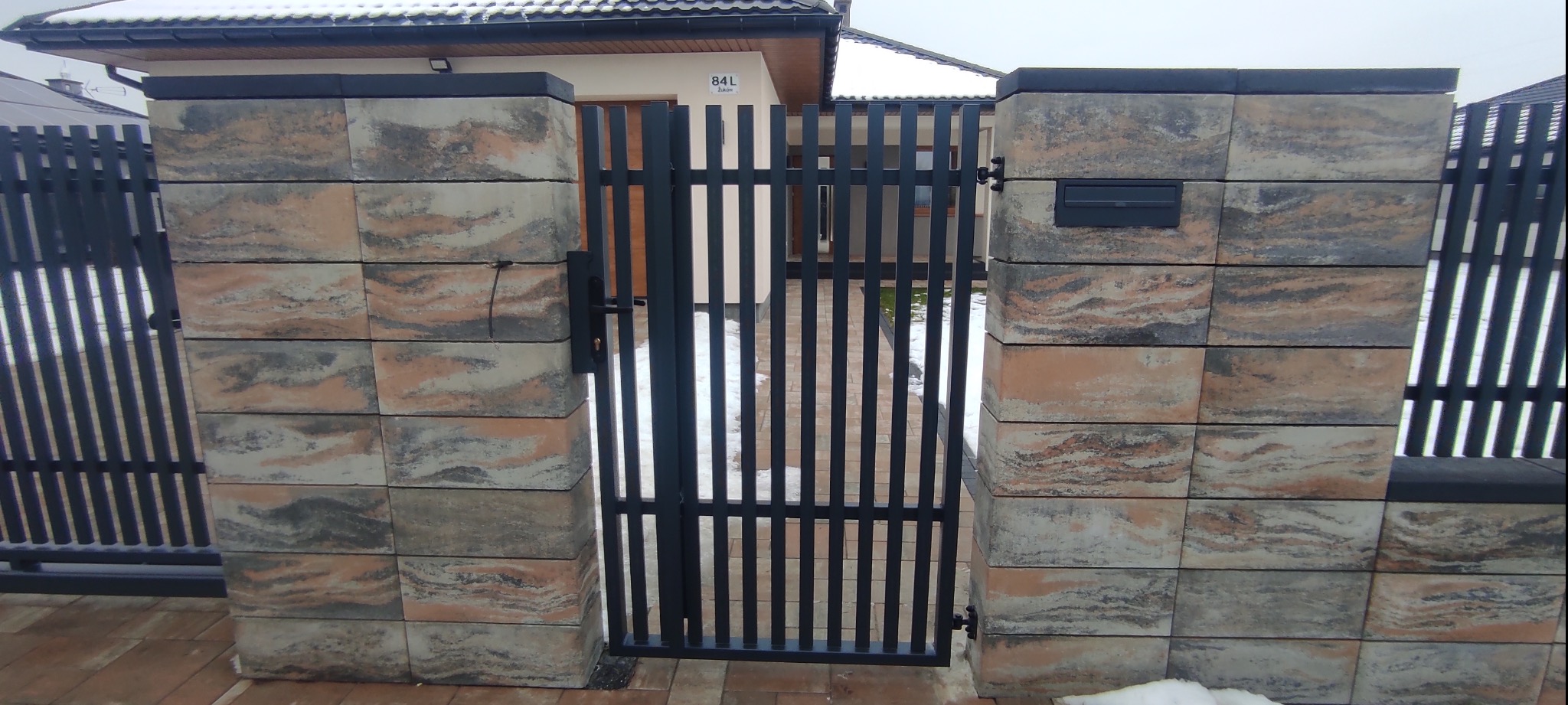 Nowoczesna brama z pionowych profili stalowych, osadzona między słupkami z dekoracyjnych bloczków betonowych w odcieniach beżu i szarości, z widocznym numerem domu na budynku w tle.