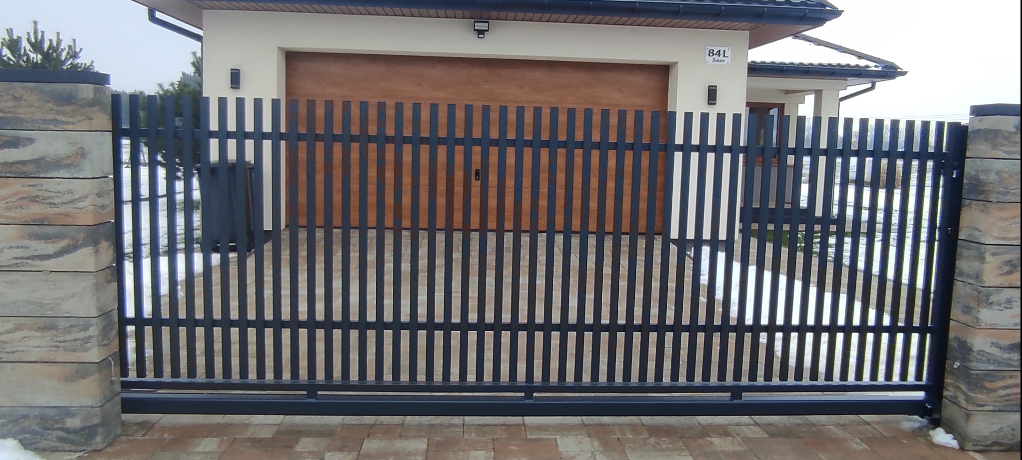 Nowoczesna, grafitowa brama wjazdowa z pionowych profili, na tle garażu z drewnianą bramą segmentową i kostki brukowej na podjeździe. Słupki bramy z dekoracyjnych bloczków betonowych.