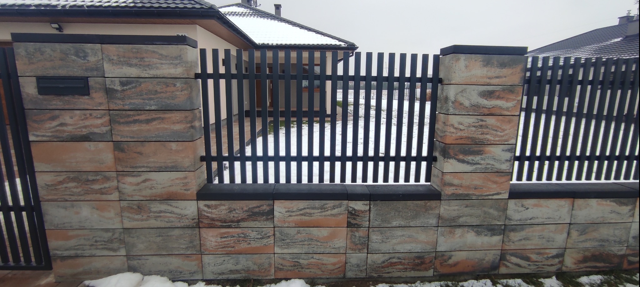 Nowoczesne ogrodzenie z metalowymi sztachetami i betonowymi słupkami o nieregularnej, marmurkowej fakturze w odcieniach szarości i beżu, z widocznym fragmentem posesji w tle.