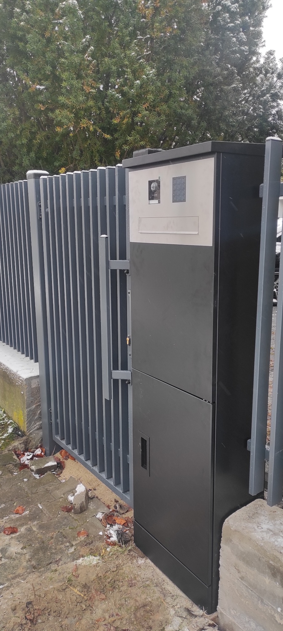 Szara brama wjazdowa z panelem domofonu i skrzynką na listy, widok z zewnątrz na tle szarego ogrodzenia panelowego i drzewa z resztkami śniegu.