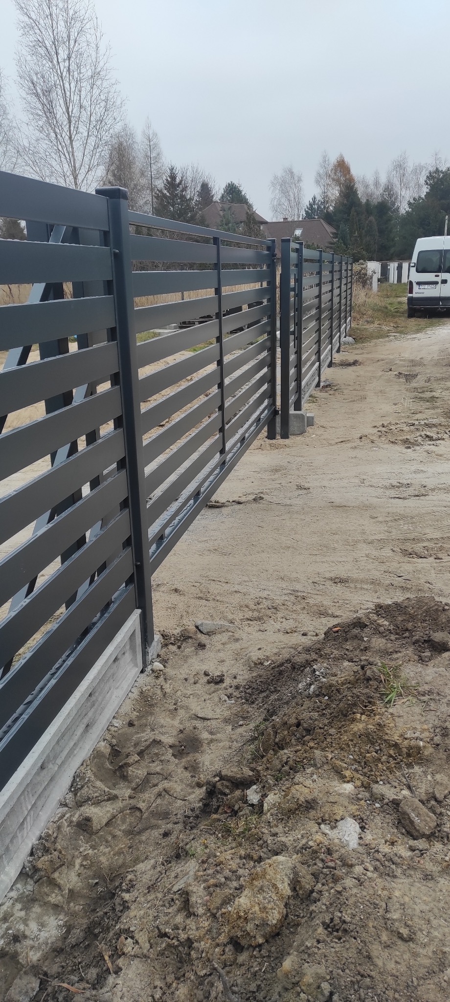 Nowoczesne, poziome, szare przęsła ogrodzeniowe na betonowej podmurówce, widoczne z boku, z samochodem dostawczym w tle.