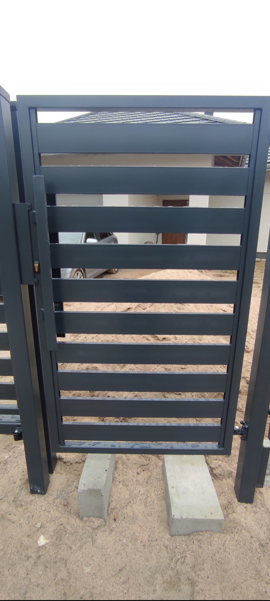 Nowoczesna, antracytowa brama wjazdowa z poziomymi profilami, tymczasowo osadzona na betonowych bloczkach, widok przez szpary na zaparkowany samochód i dom z szarą dachówką.