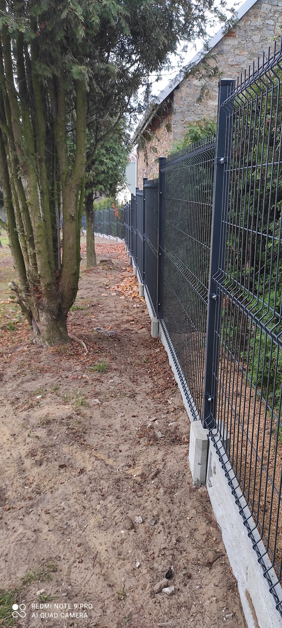 Długa, czarna konstrukcja z paneli ogrodzeniowych, zamontowana na betonowej podmurówce, oddziela posesję porośniętą drzewami od sąsiedniego budynku.