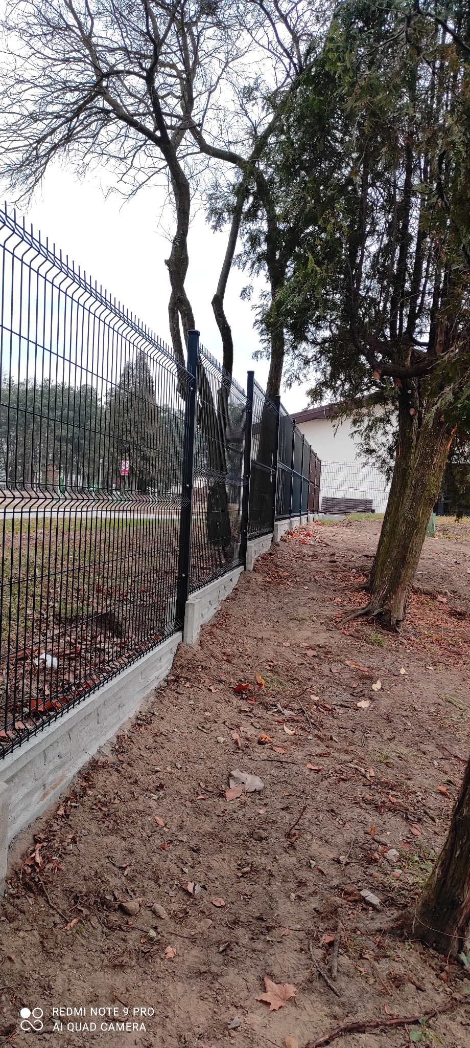 Czarne panele ogrodzeniowe na betonowej podmurówce, widok z boku wzdłuż ogrodzenia, z drzewami w tle i opadłymi liśćmi na ziemi.