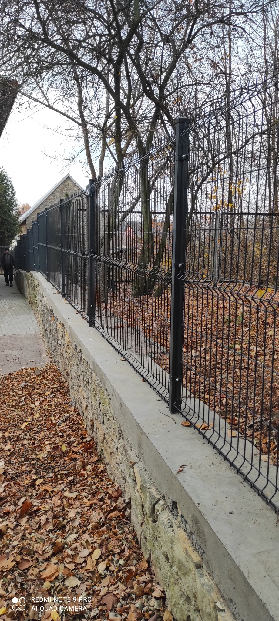 Czarne, nowoczesne ogrodzenie z paneli na betonowo-kamiennym murku, jesienne liście na ziemi, widok z boku.