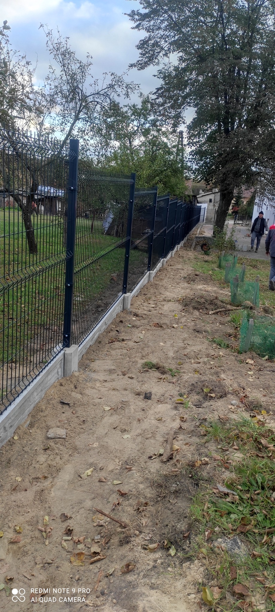 Zamontowane ciemne ogrodzenie panelowe na betonowej podmurówce, widoczne zabezpieczone siatką sadzonki drzew, w tle budynek mieszkalny i spacerujący mężczyźni.