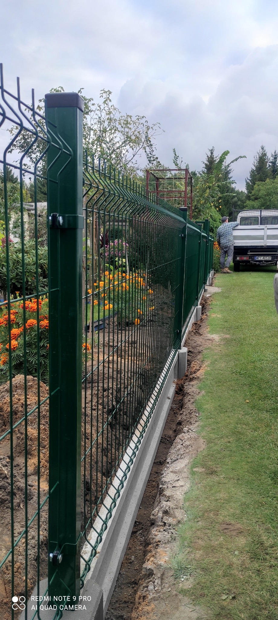 Zielone panele ogrodzeniowe na betonowej podmurówce w trakcie montażu, widoczny częściowo samochód dostawczy i pracownik w tle.