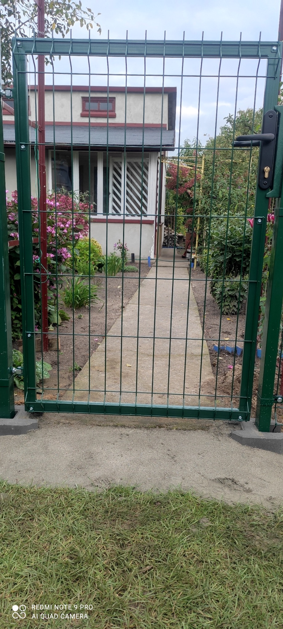 Zielona brama z paneli ogrodzeniowych z widokiem na ścieżkę prowadzącą do domu z ogrodem.