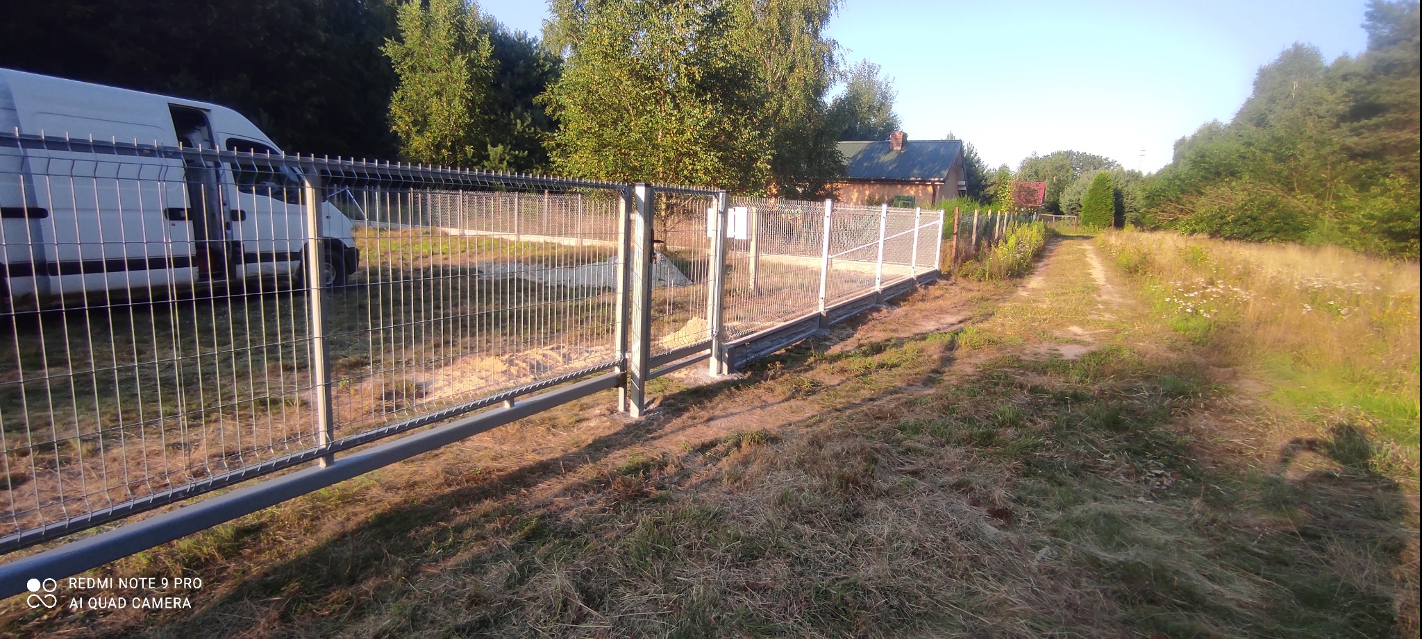 Nowo zamontowane, ocynkowane przęsła z siatki zgrzewanej, tworzące ogrodzenie posesji na wsi, z widocznym busem dostawczym w tle.