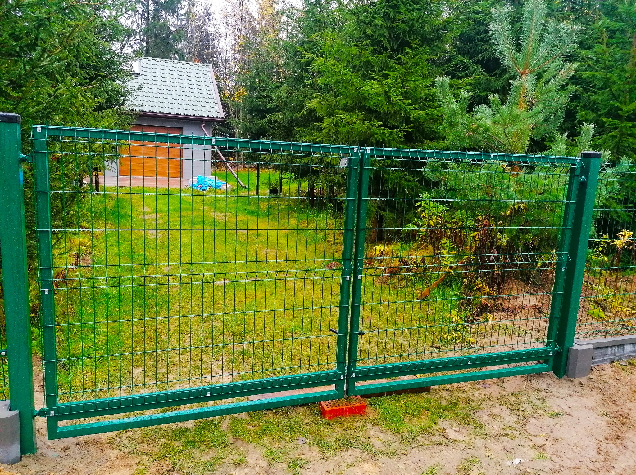Zielona, dwuskrzydłowa brama z paneli ogrodzeniowych na tle trawnika i domku gospodarczego, tymczasowo podparta cegłą.