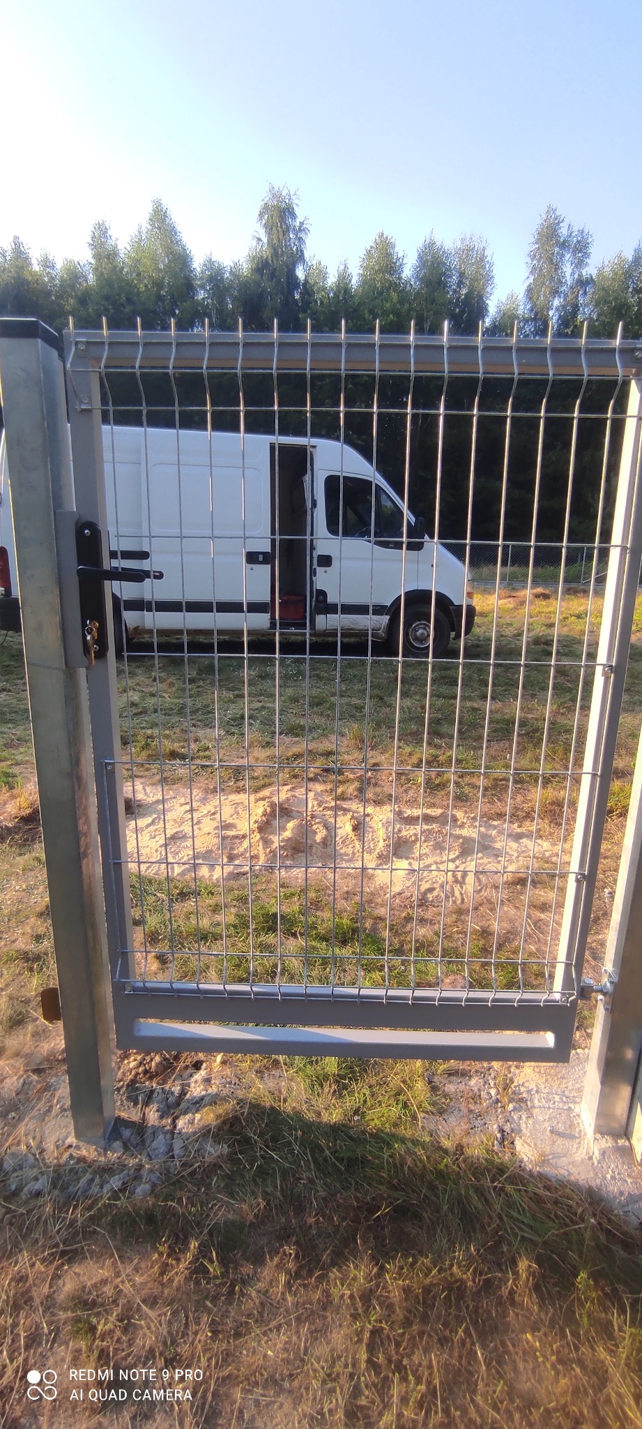 Szara brama panelowa z widocznym zamkiem i kluczem, w tle biały samochód dostawczy z otwartymi drzwiami na tle drzew.