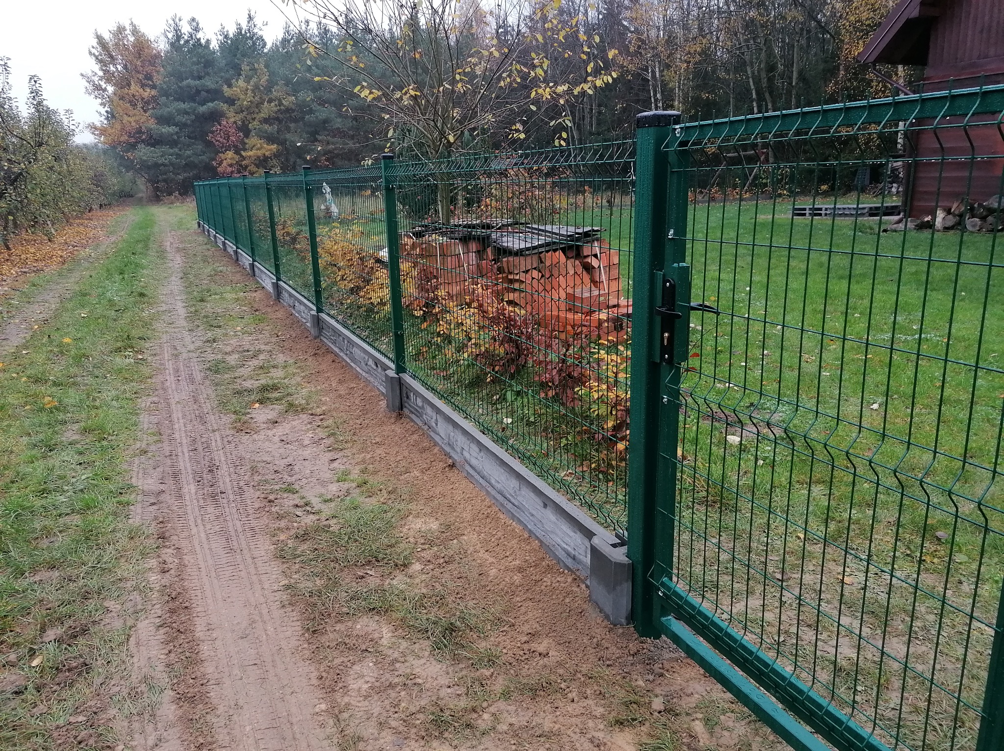 Długi, zielony system ogrodzeniowy z paneli z podmurówką betonową, oddzielający trawnik od polnej drogi, z stosem drewna opałowego widocznym przez siatkę.