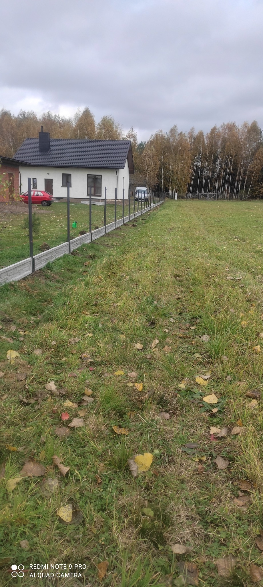 Montaż betonowej podmurówki z metalowymi słupkami na działce z domem jednorodzinnym i jesiennym drzewostanem w tle.