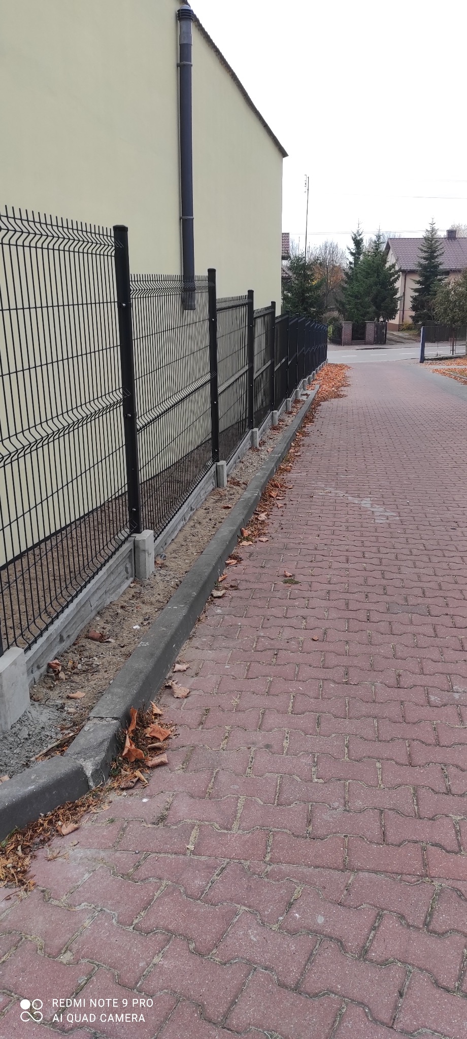 Czarny, metalowy płot panelowy z betonową podmurówką, zamontowany w Krzczonowie wzdłuż chodnika z czerwonej kostki, opadłe liście na chodniku i przy krawężniku.