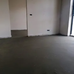 Surowy, szary jastrych w nowym budynku z widocznymi puszkami elektrycznymi w ścianach, przed oknem widoczne drzewa.