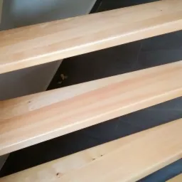 Jasne, drewniane stopnie schodów z widocznymi słojami drewna i małymi sękami, ułożone nad ciemną, prawdopodobnie kamienną podłogą, z fragmentem ściany w tle.