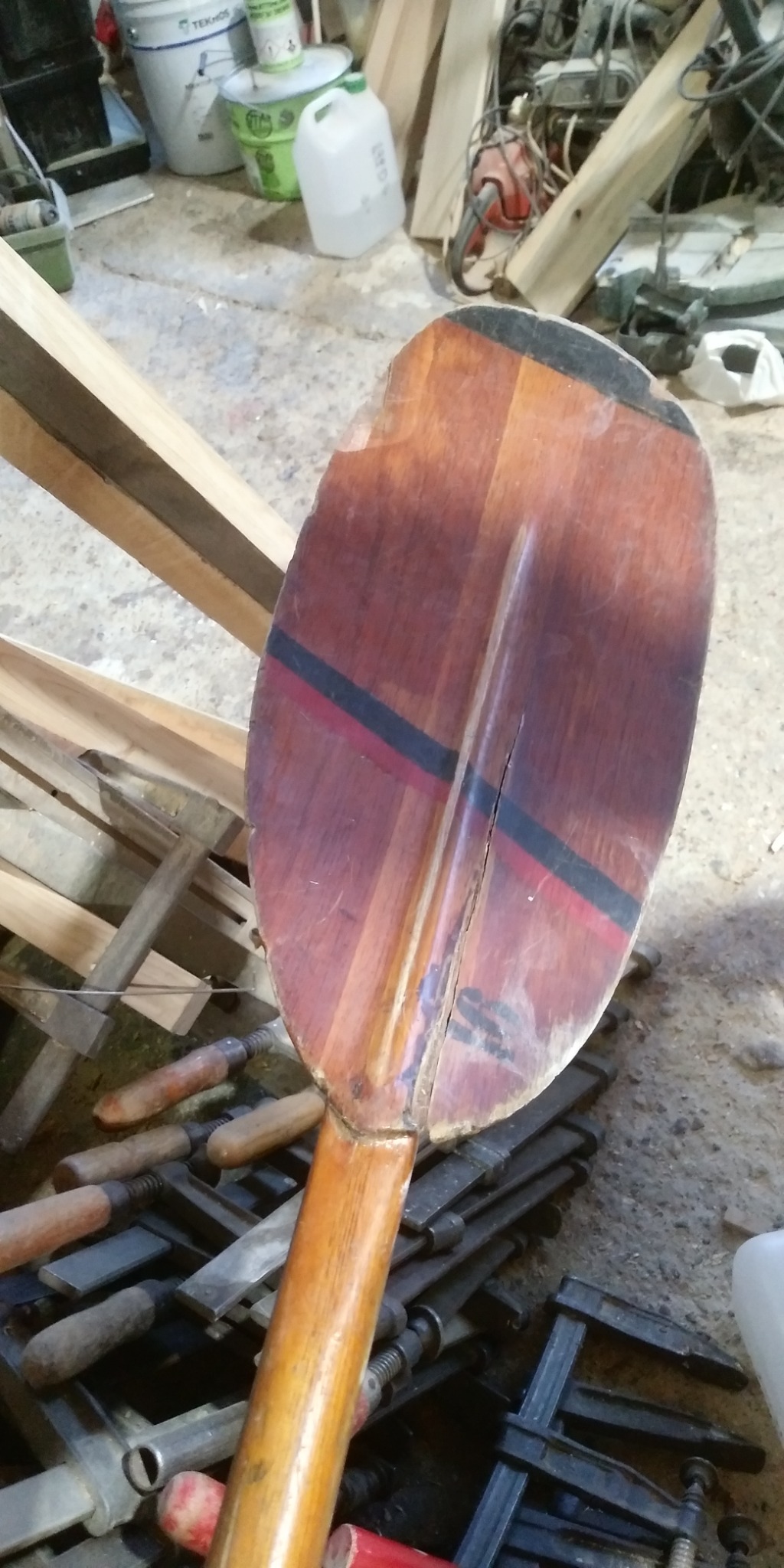 Drewniane wiosło z pęknięciem na piórze, leżące na stercie ścisków stolarskich w warsztacie.