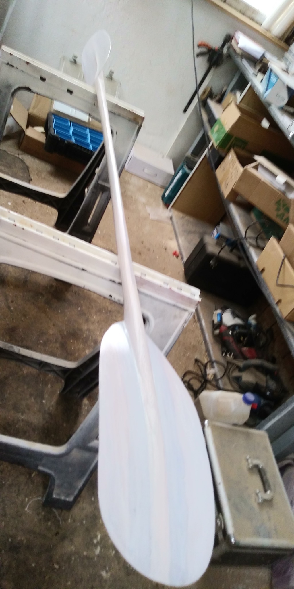 Pomalowana na biało drewniana łopata stoi oparta o stół warsztatowy w zagraconym pomieszczeniu, widoczne narzędzia i materiały budowlane w tle.