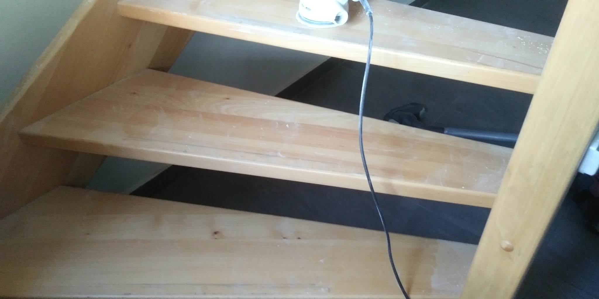 Drewniane schody podczas renowacji; widoczne ślady szlifowania i szlifierka oscylacyjna na górnym stopniu.