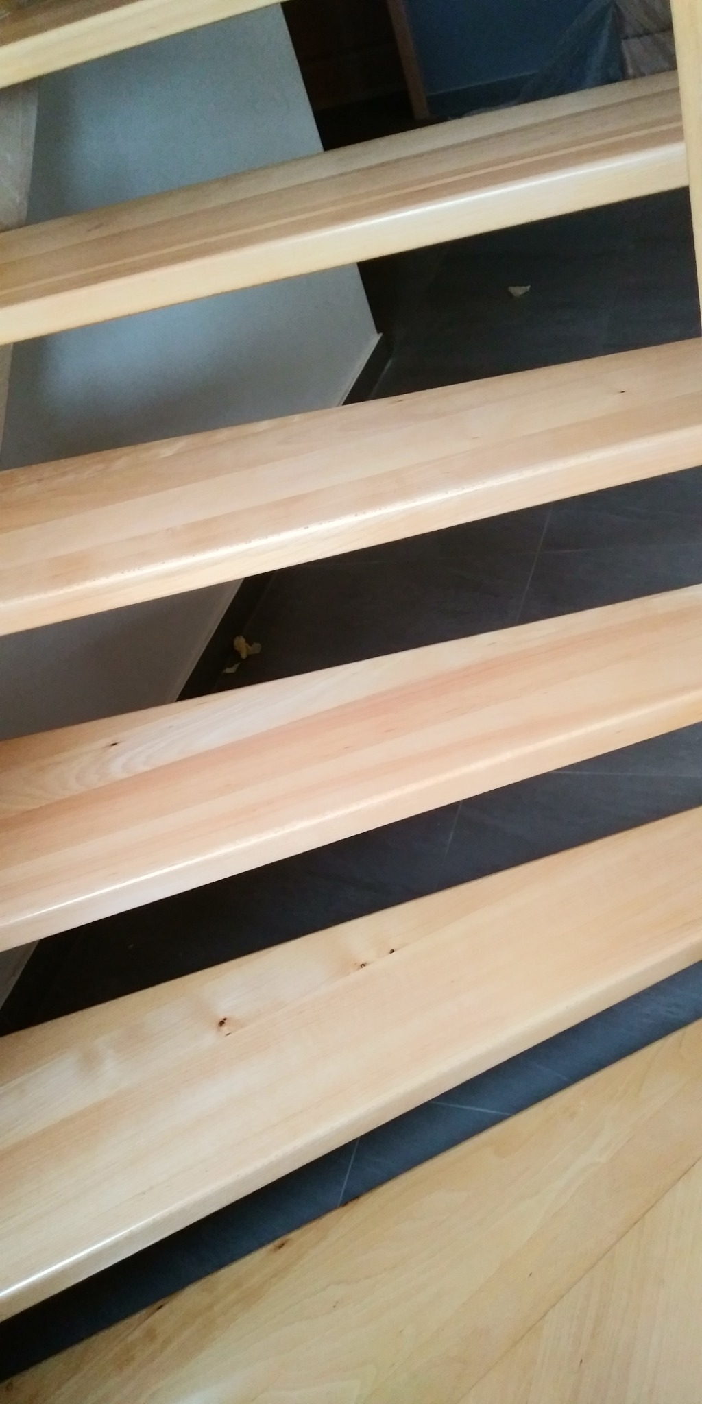 Jasne, drewniane stopnie schodów z widocznymi słojami drewna i małymi sękami, ułożone nad ciemną, prawdopodobnie kamienną podłogą, z fragmentem ściany w tle.