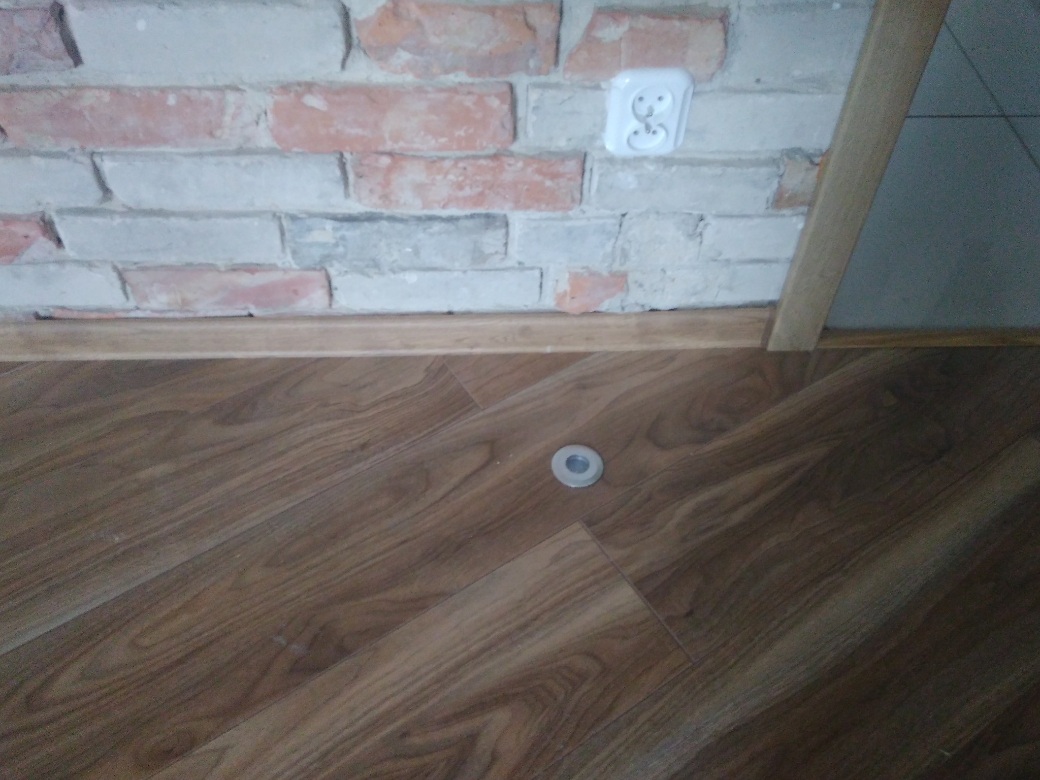 Fragment wnętrza z ułożoną podłogą z paneli laminowanych w odcieniach brązu, widoczna ceglana ściana z gniazdkiem elektrycznym i fragmentem listwy przypodłogowej oraz element wentylacyjny w podłodze.