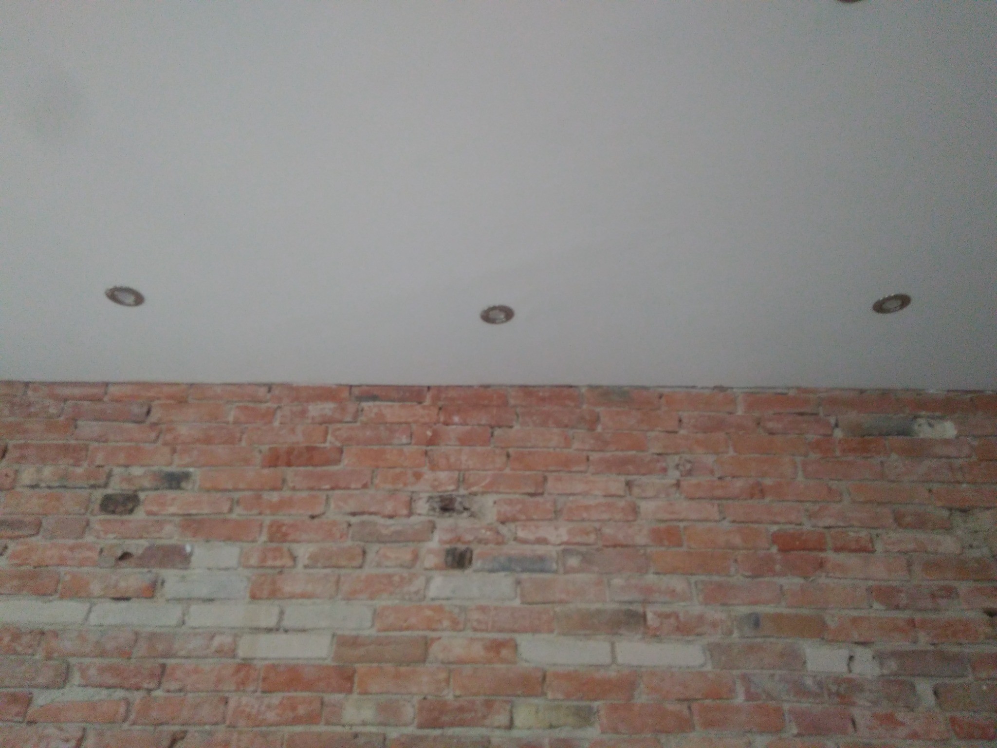 Biały sufit z trzema okrągłymi lampami halogenowymi nad ścianą z czerwonej cegły z widocznymi spoinami.