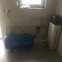 Niebieski zbiornik hydroforowy i zmiękczacz wody w kotłowni wyłożonej płytkami ceramicznymi, widoczne rury instalacji wodnej i okno z parapetem.